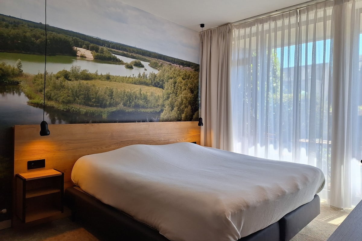 A bedroom with a large bed, a nightstand, a wall-mounted lamp, and a view of a lake through sheer curtains.