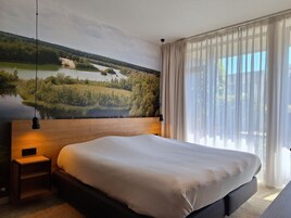 Quarto com uma cama grande, mesa de cabeceira, luminária na parede e vista para um lago através de cortinas translúcidas.