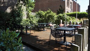 An outdoor patio with tables and chairs, surrounded by greenery and a brick building in the background.