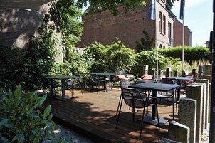 Un patio al aire libre con mesas y sillas, rodeado de follaje y un edificio de ladrillo al fondo.
