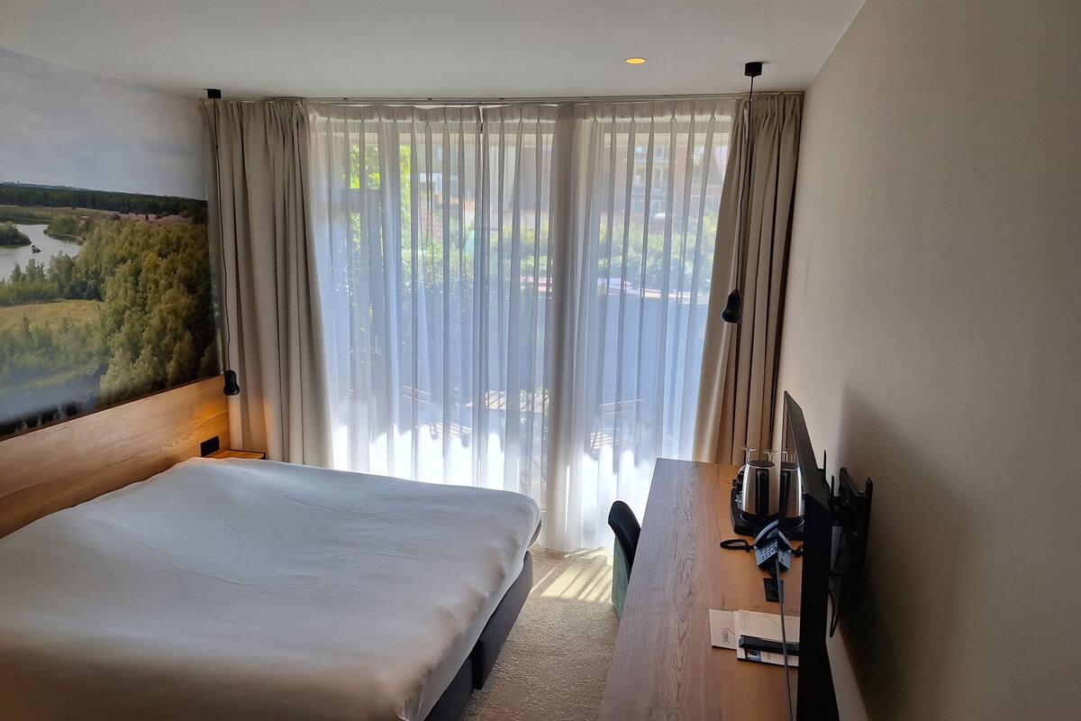 A hotel room with a large bed, a wooden desk with a telephone and a coffee maker, and a view of greenery through sheer curtains.