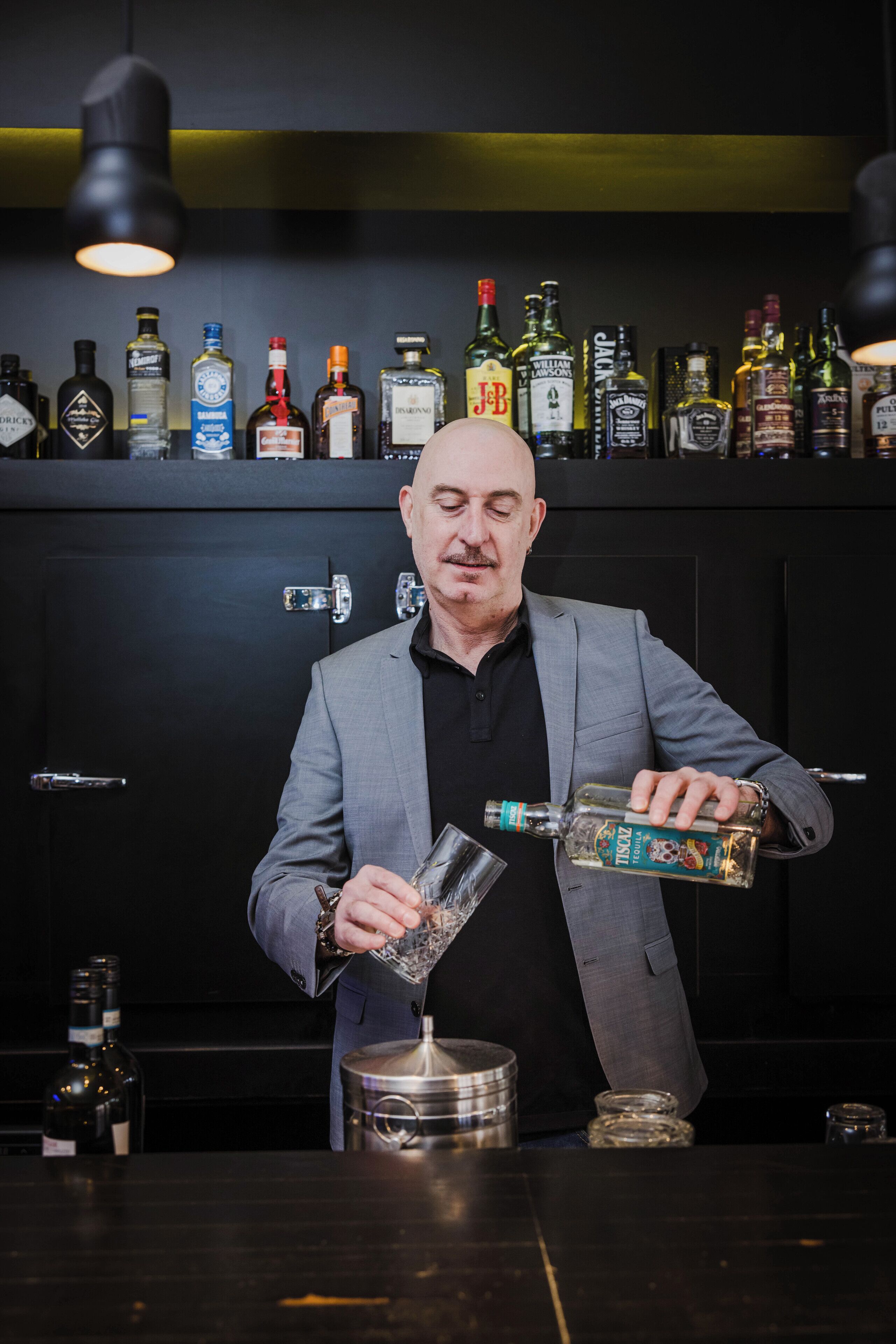 A man in a suit pouring a drink into a glass at a bar with a variety of liquor bottles in the background.