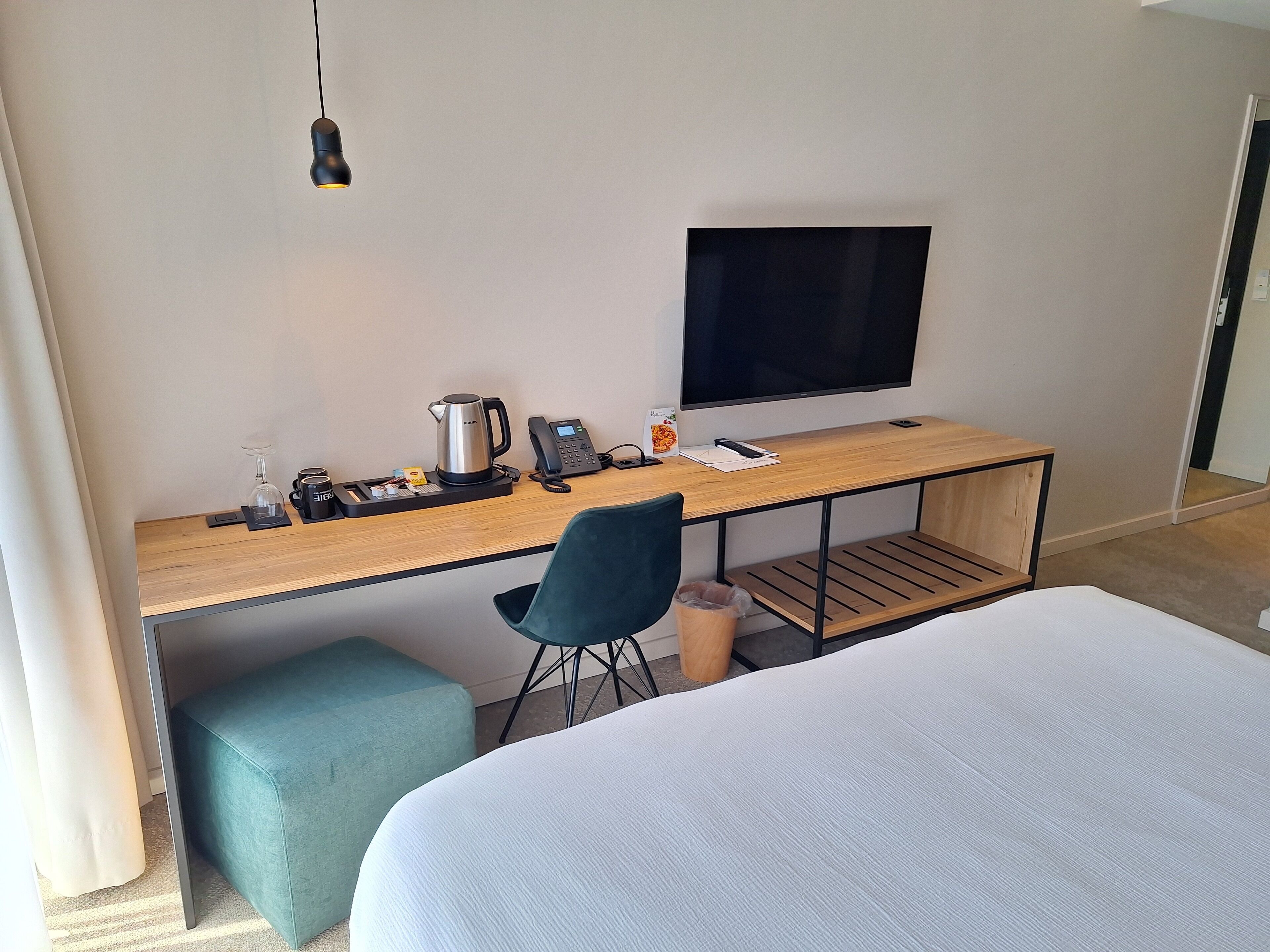 Quarto de hotel com cama, escrivaninha, computador, televisão, máquina de café e lixeira.