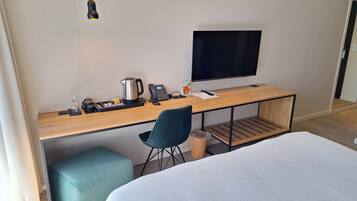 Quarto de hotel com cama, escrivaninha, computador, televisão, máquina de café e lixeira.