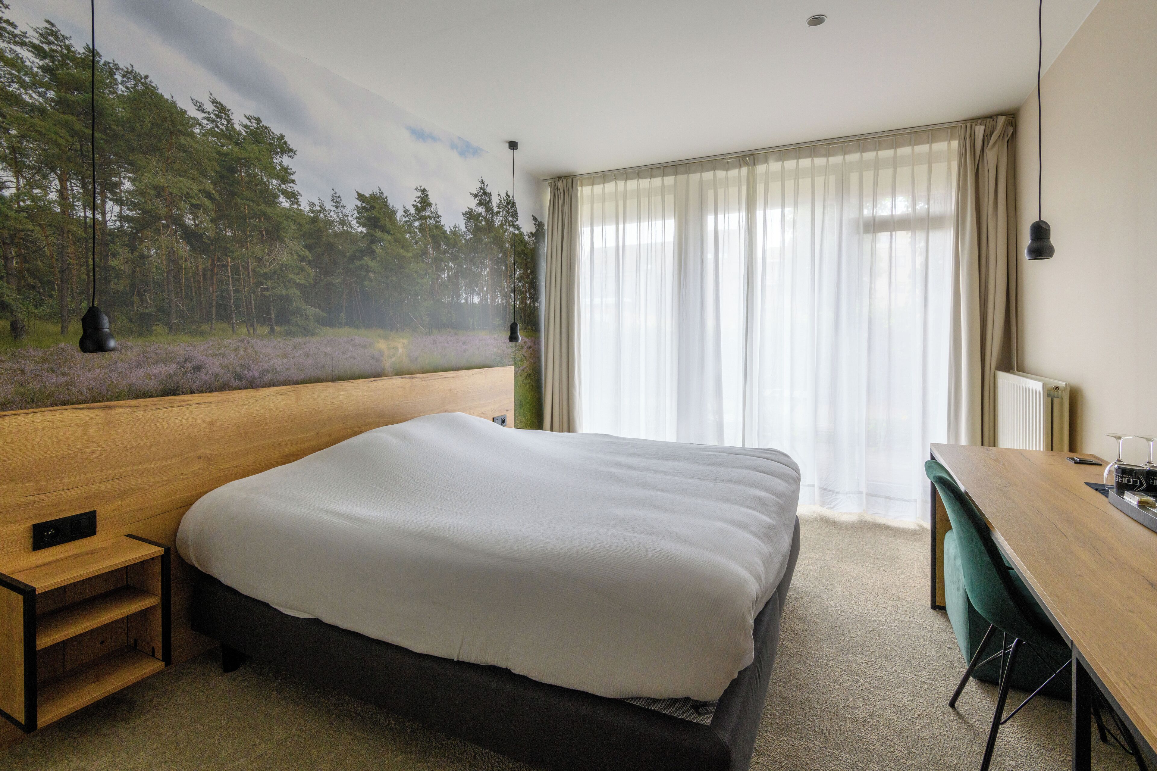 Une chambre à coucher avec un grand lit, un bureau en bois et une vue sur une forêt, visible à travers de fines rideaux.