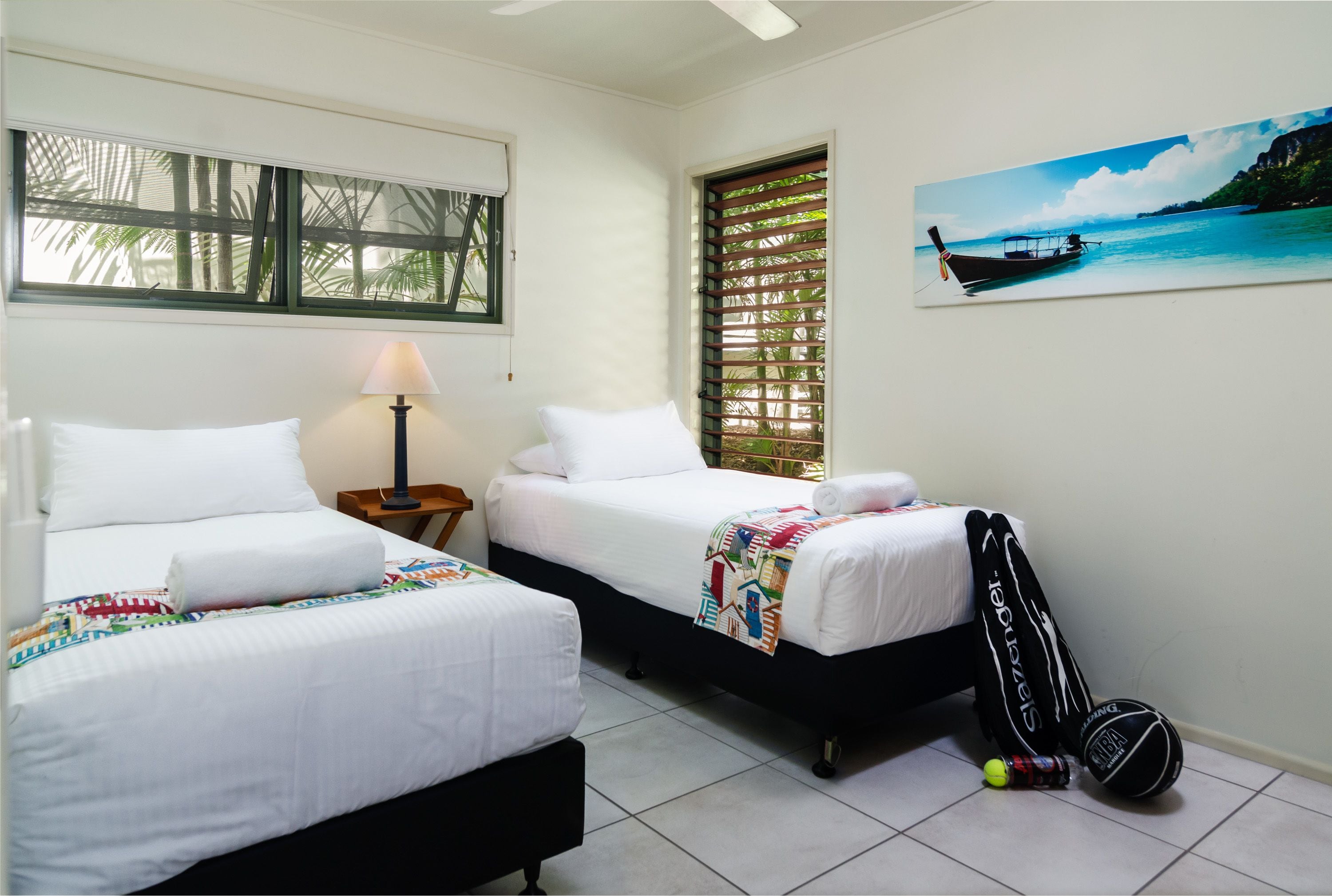 A hotel room with two beds, a window with blinds, a lamp, a tennis racket, and a sports ball on the floor.