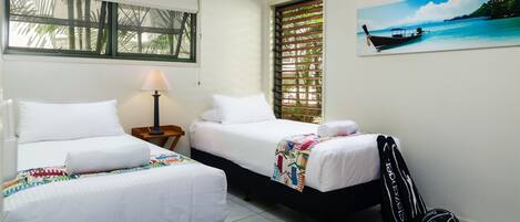 A hotel room with two beds, a window with blinds, a lamp, a tennis racket, and a sports ball on the floor.