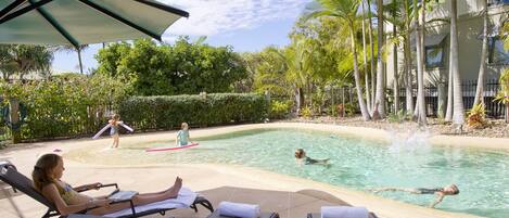 Un área de piscina con sillones de descanso, sombrillas y una piscina donde gente nada y juega.