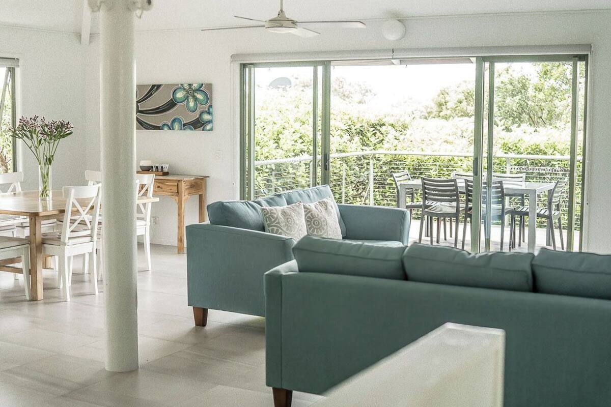 A modern living room with a teal sofa, a dining area with white chairs, and a balcony with outdoor seating.