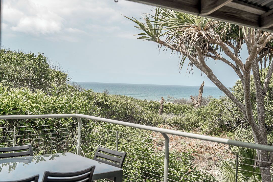 Un balcón con mesa y sillas, con vista a un paisaje costero con vegetación y el mar.
