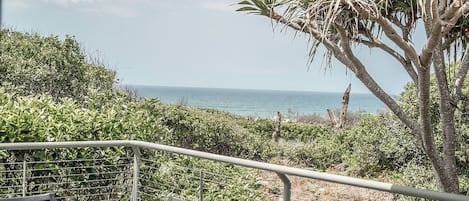 Un balcón con mesa y sillas, con vista a un paisaje costero con vegetación y el mar.