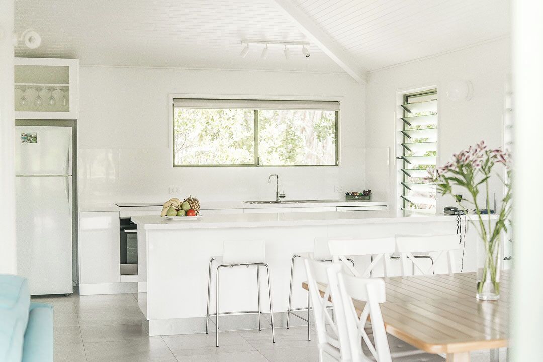 Una cocina moderna con armarios blancos, una isla central con taburetes, una ventana con vistas al verdor y una zona de comedor con mesa de madera.