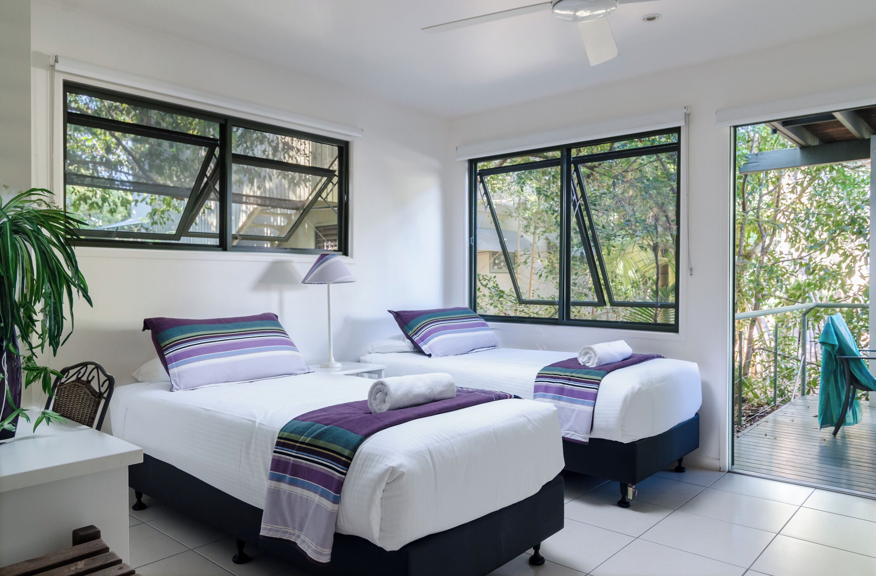 A room with two beds, a desk, and a chair. There are large windows with views of trees and a balcony.