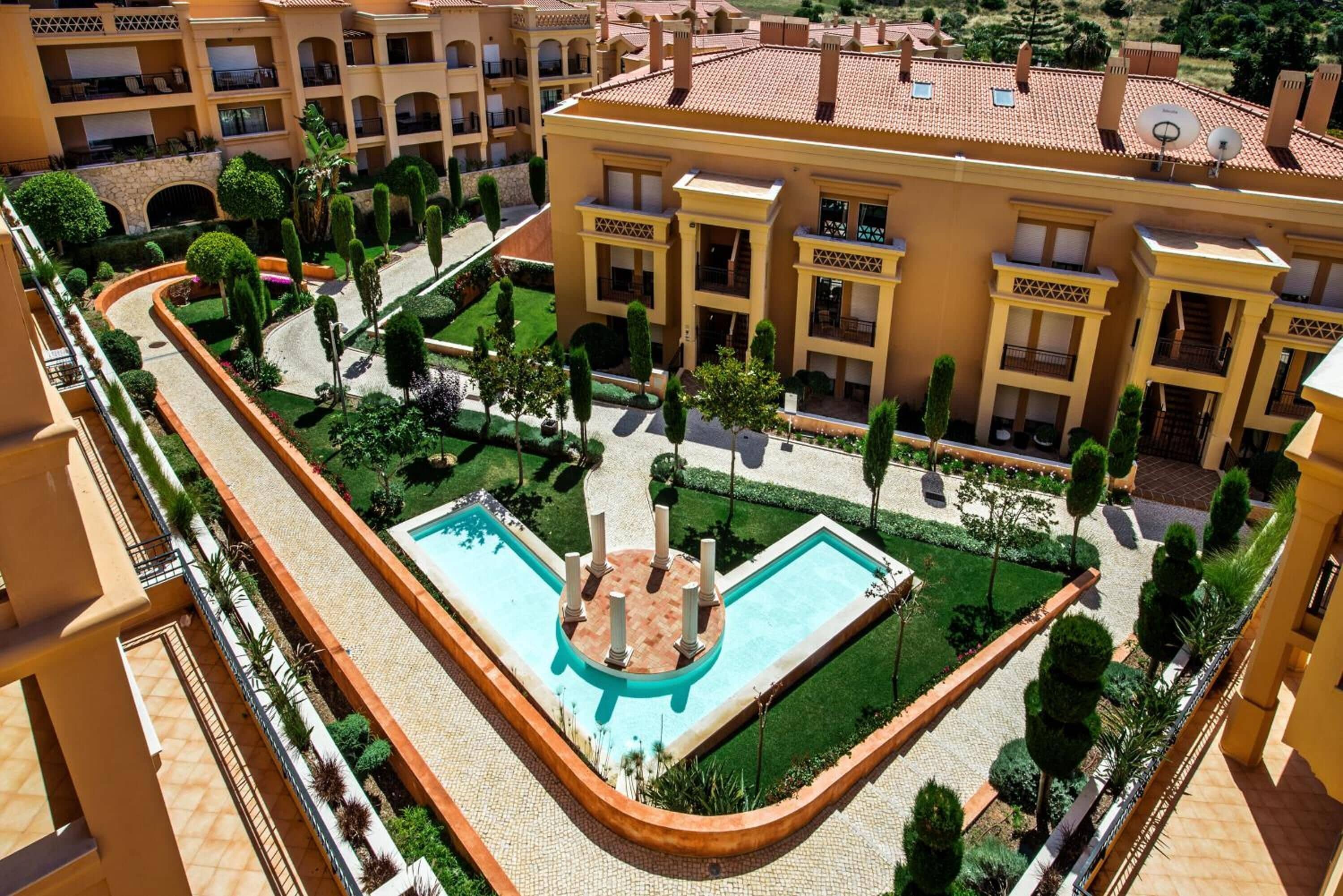 Une cour d’hôtel avec une piscine centrale, entourée de haies soigneusement taillées et de diverses espèces d’arbres.