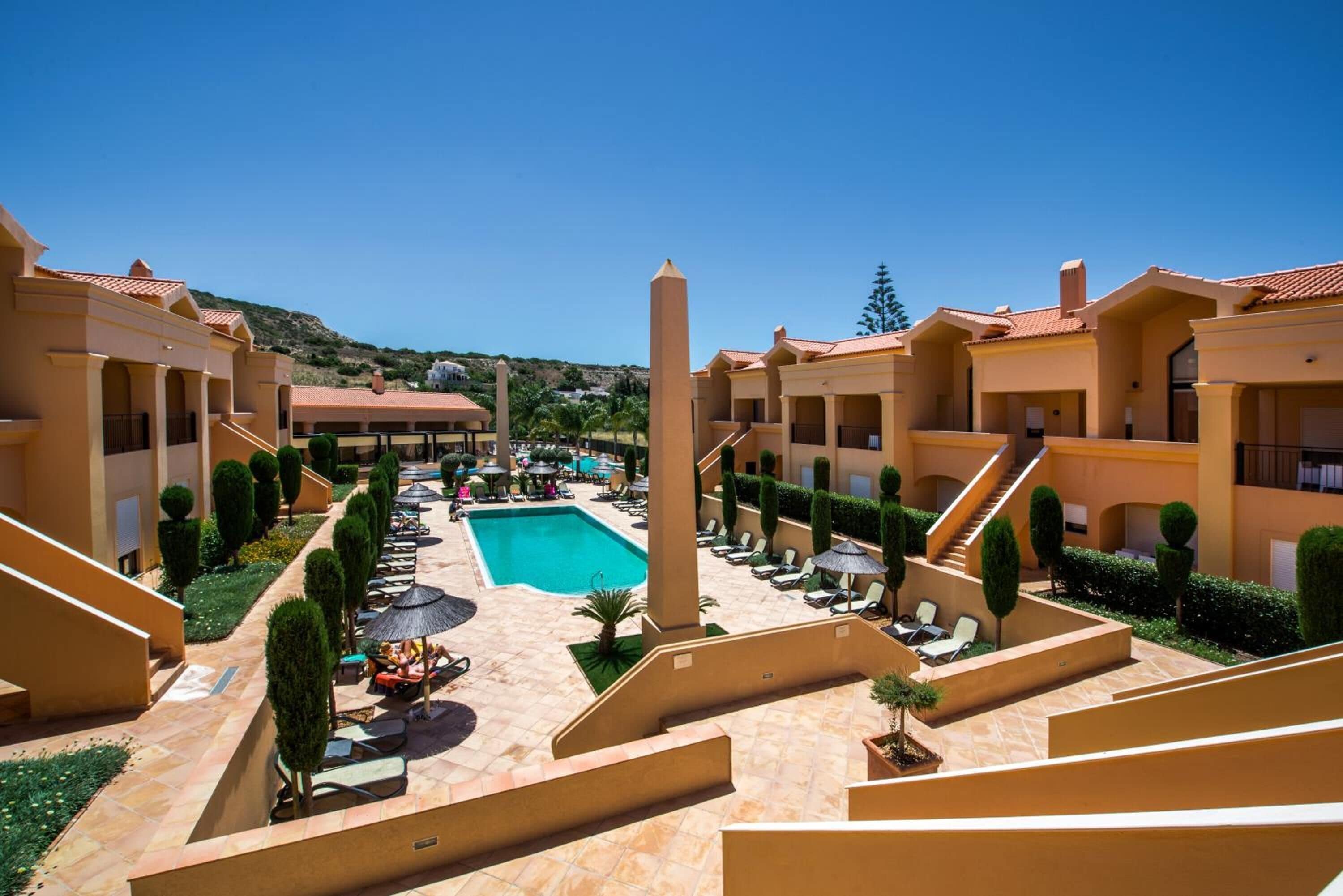 A hotel courtyard with a swimming pool, sun loungers, and a central obelisk.