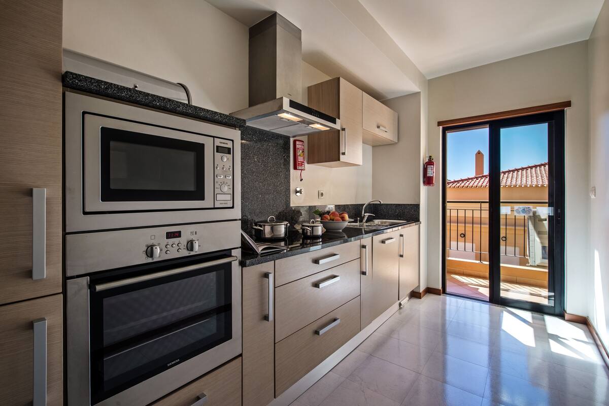 A modern kitchen with stainless steel appliances, wooden cabinets, and a large window overlooking a balcony.
