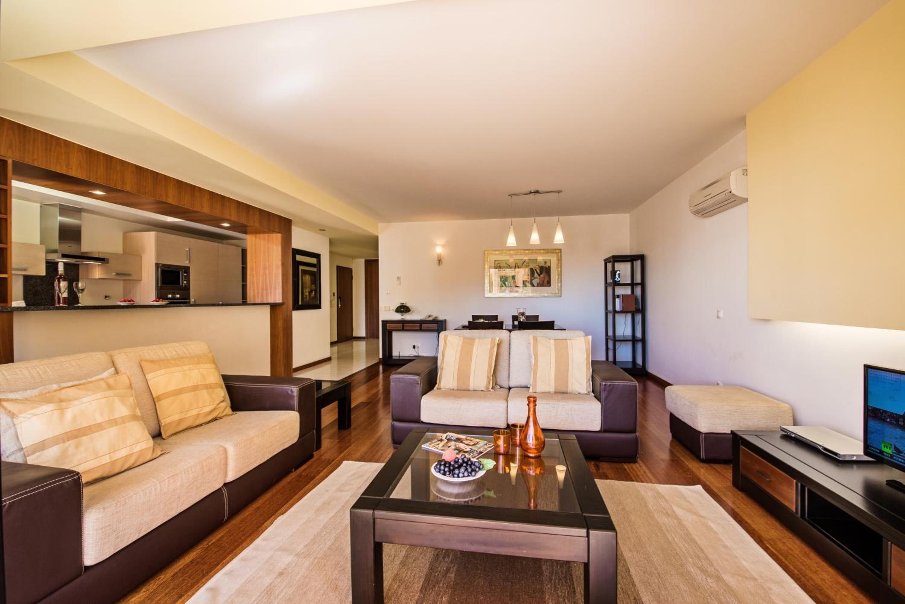 A modern living room with a sofa, coffee table, and a television. There is a kitchen area visible in the background.