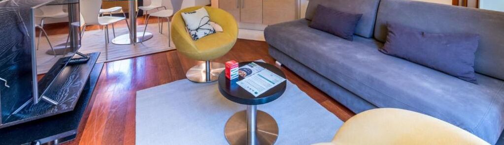 Un salon moderne avec un canapé gris, un fauteuil jaune, une petite table ronde sur laquelle se trouve un magazine, et un téléviseur posé sur un plancher en bois.