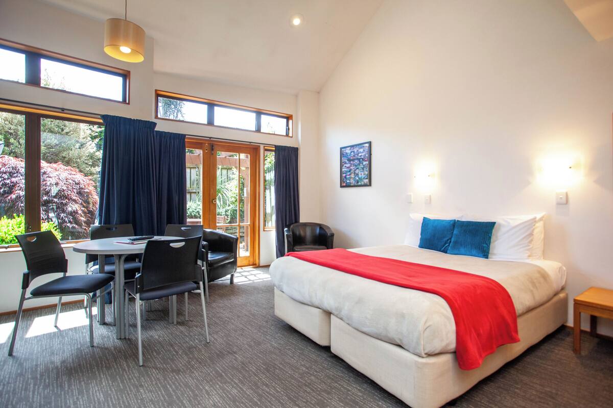 Une chambre d’hôtel comprenant un lit, un bureau avec des chaises, une petite table et une fenêtre avec des rideaux.