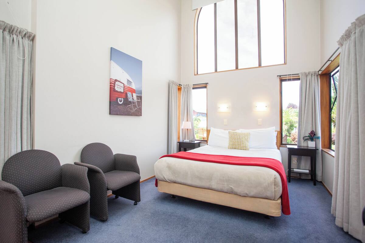 Une chambre d’hôtel avec un grand lit, deux fauteuils, une table de chevet, une lampe et une fenêtre avec des rideaux.