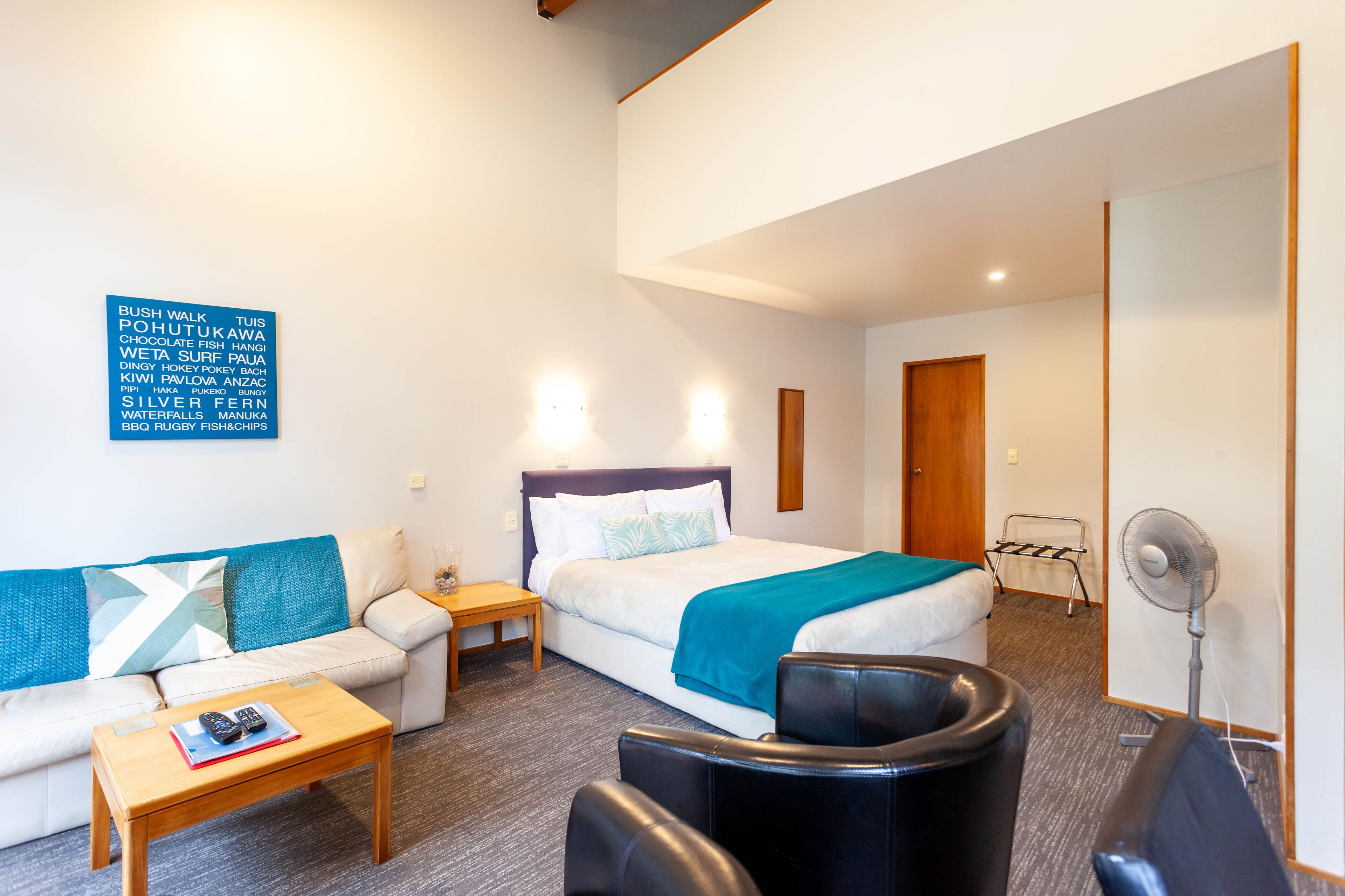 A hotel room with a bed, a sofa, a small table, a chair, a standing fan, and a wall-mounted light fixture.