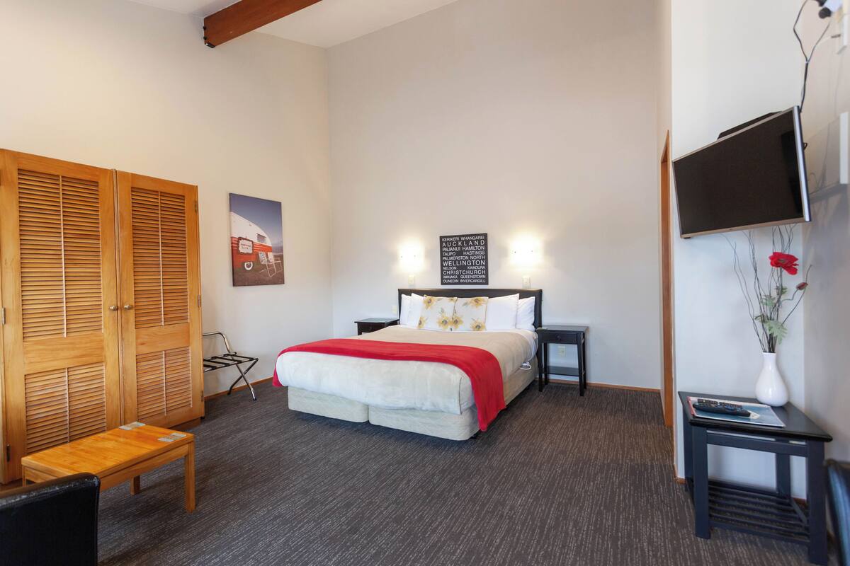Une chambre d’hôtel comprenant un lit, une armoire en bois, une petite table, une chaise, un téléviseur fixé au mur et un luminaire fixé au mur.