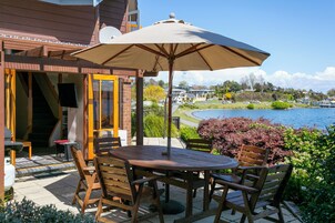 Un patio al aire libre con una mesa y sillas de madera, un gran paraguas y vistas a un lago y un pueblo al fondo.
