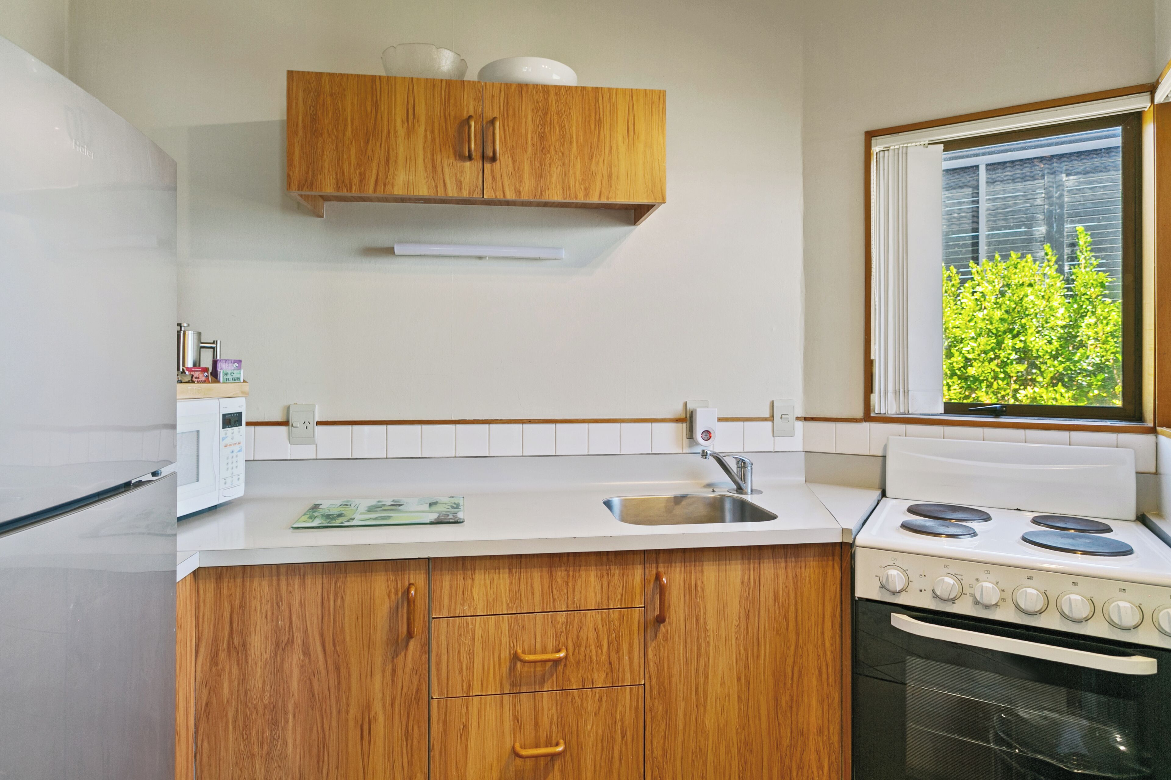 Cocina compacta con armarios de madera, encimera blanca, estufa y ventana con vistas a la vegetación.
