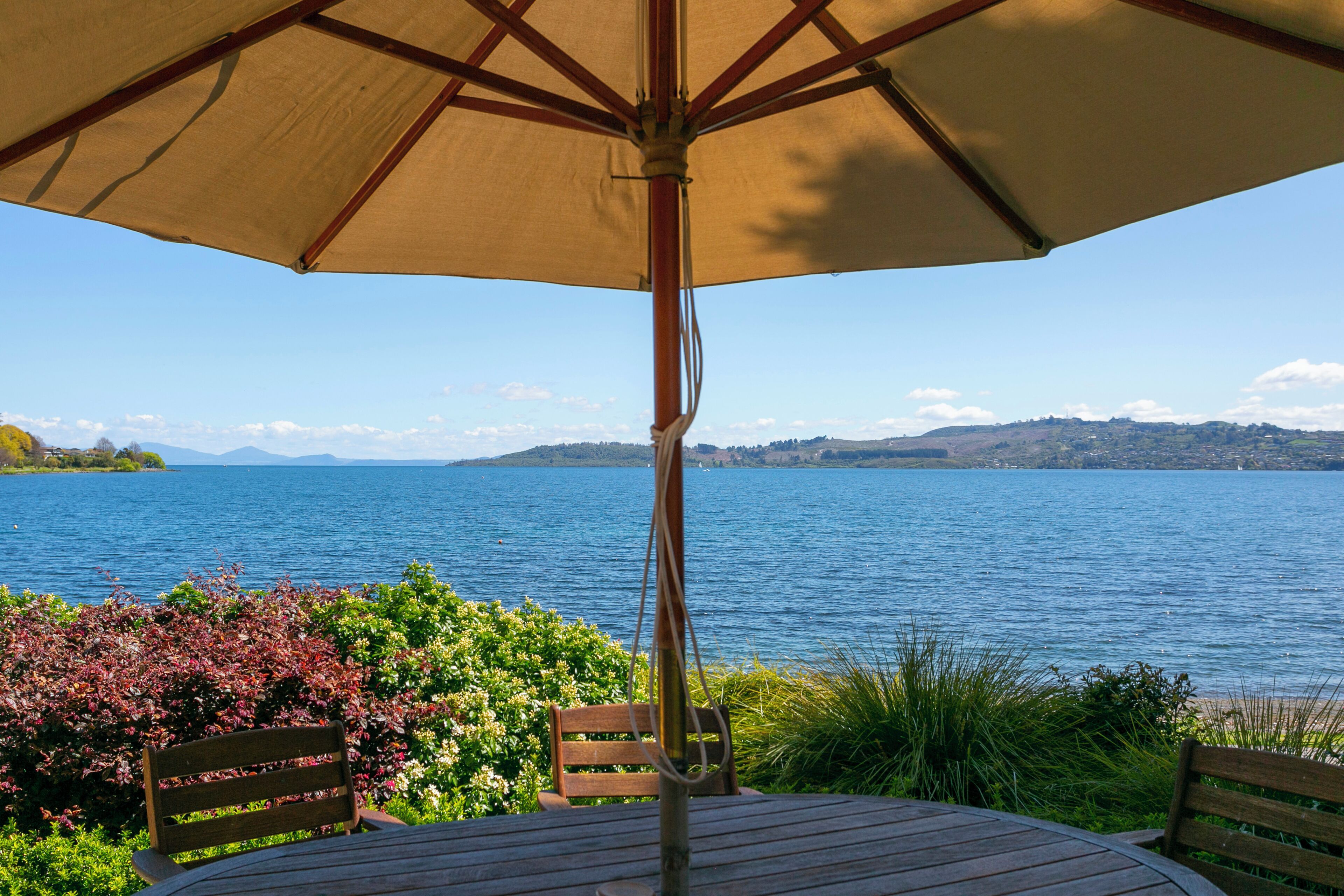 A table and chairs under a large umbrella overlooking a lake with a view of distant mountains.