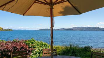 A table and chairs under a large umbrella overlooking a lake with a view of distant mountains.