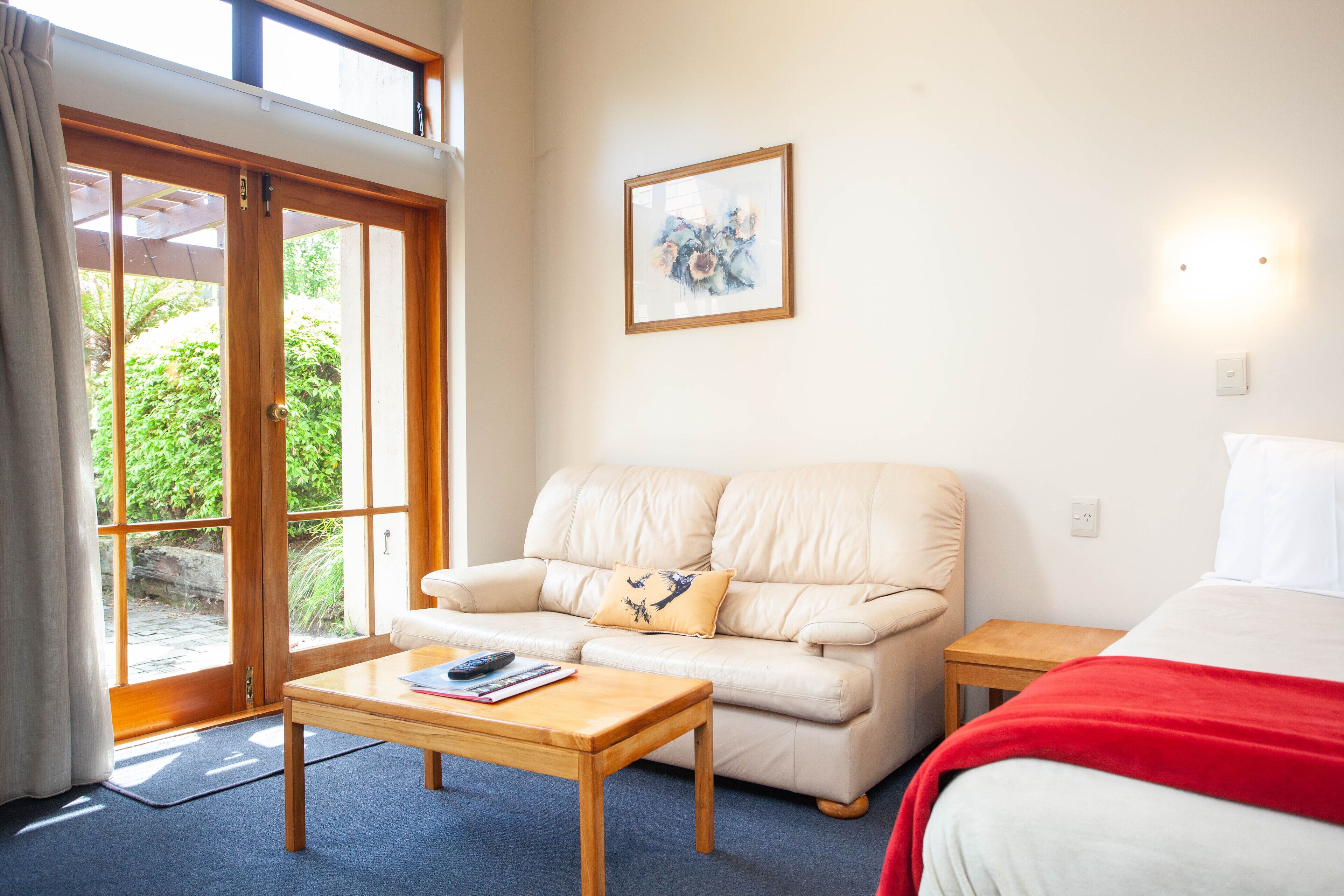 Una habitación con un sofá color crema, una mesa de centro de madera, una cama con una colcha roja, un cuadro en la pared y vistas a la vegetación por la puerta.