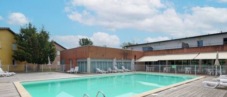 A rectangular swimming pool with a wooden deck, surrounded by lounge chairs and umbrellas, with buildings in the background.