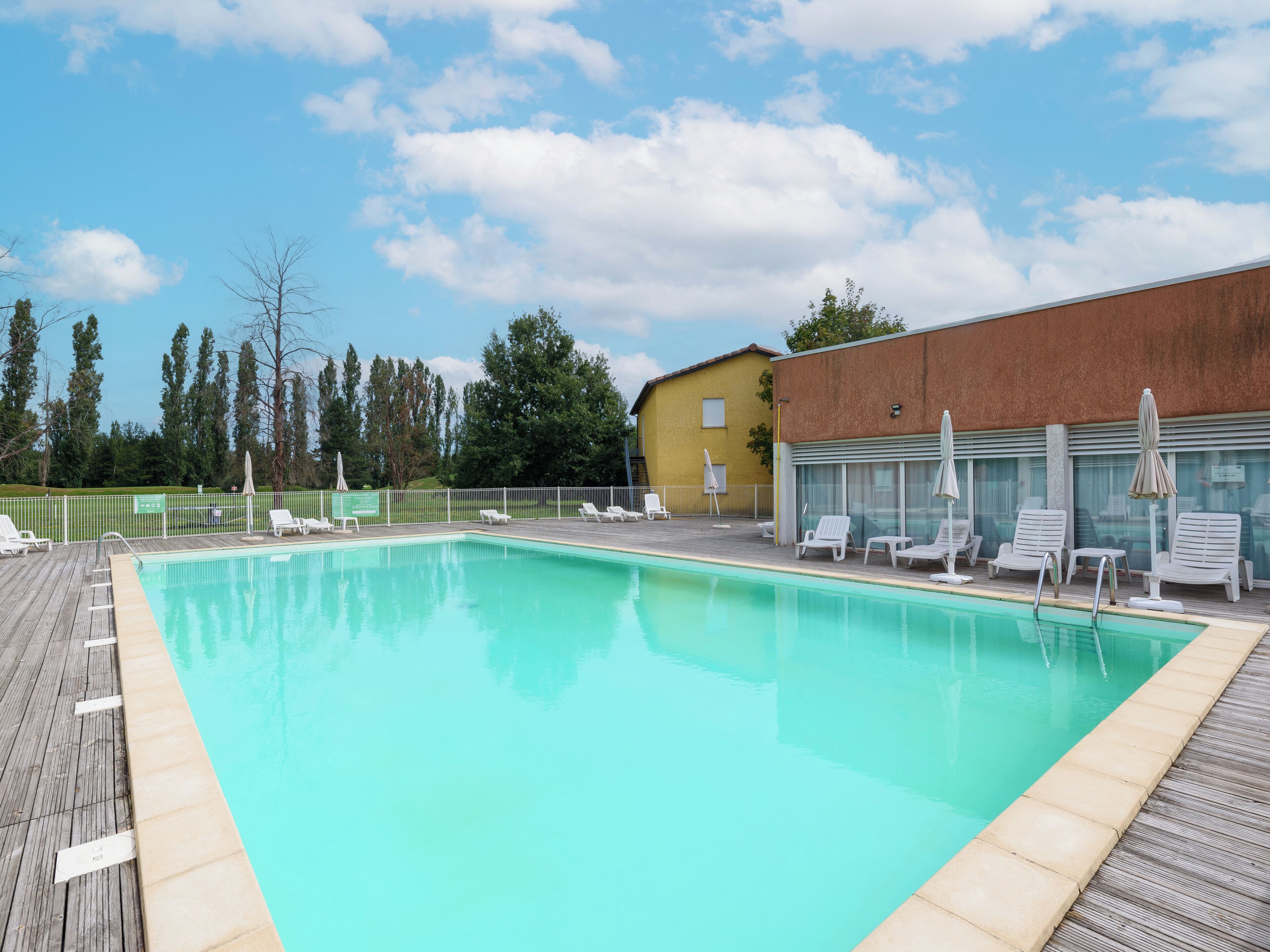 A swimming pool with lounge chairs and umbrellas, surrounded by a wooden deck and a building with a yellow facade.