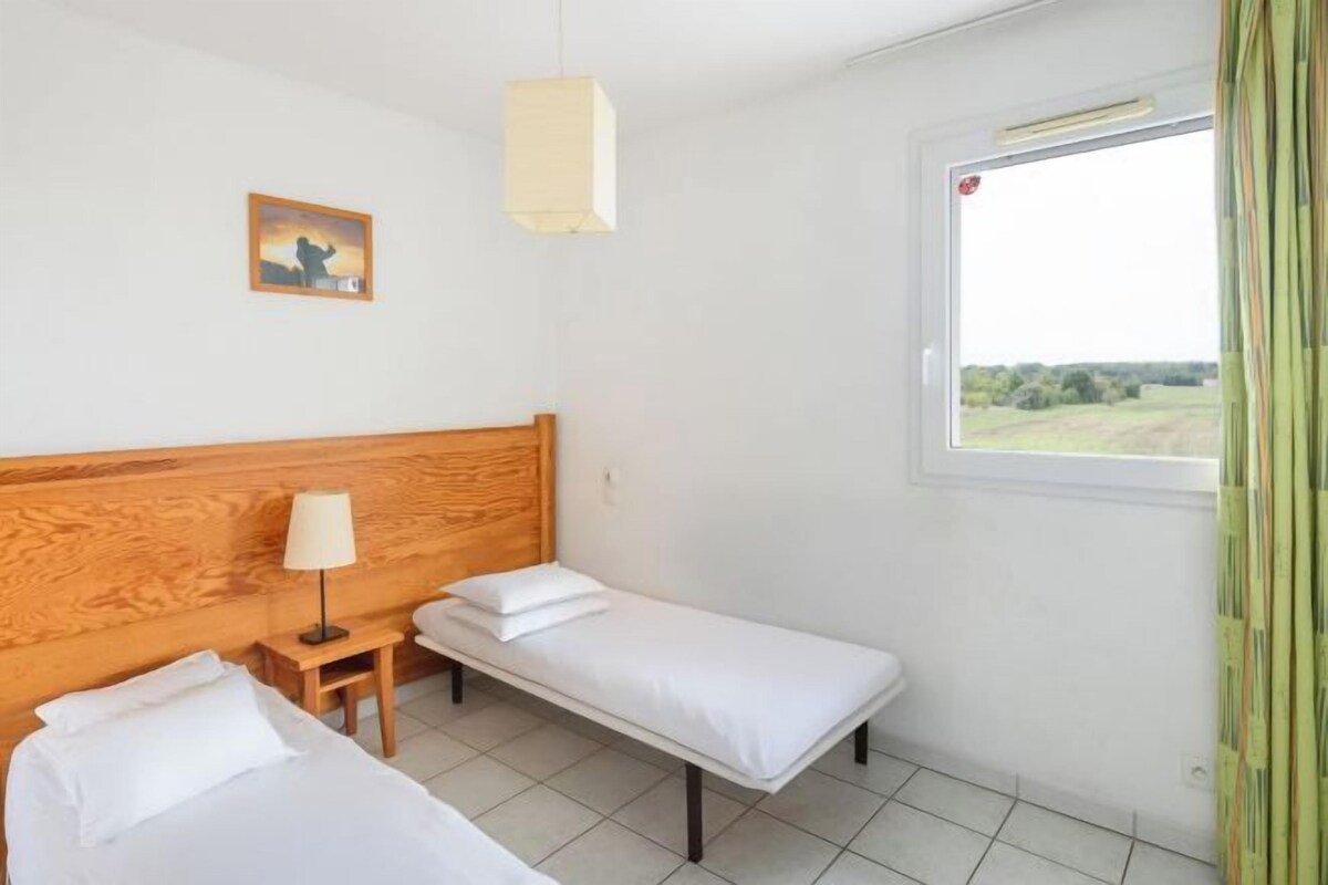 Une chambre avec deux lits simples, une tête de lit en bois, une petite table de chevet avec une lampe, une fenêtre donnant sur la campagne et un tableau au mur.