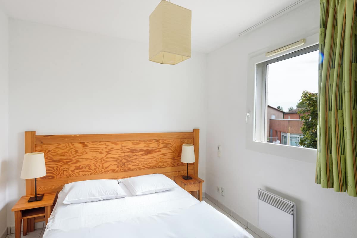 Hotelkamer met een houten hoofdeinde, twee nachtkastjes met lampen, een groot bed met witte lakens, een raam met gordijnen en een radiator.