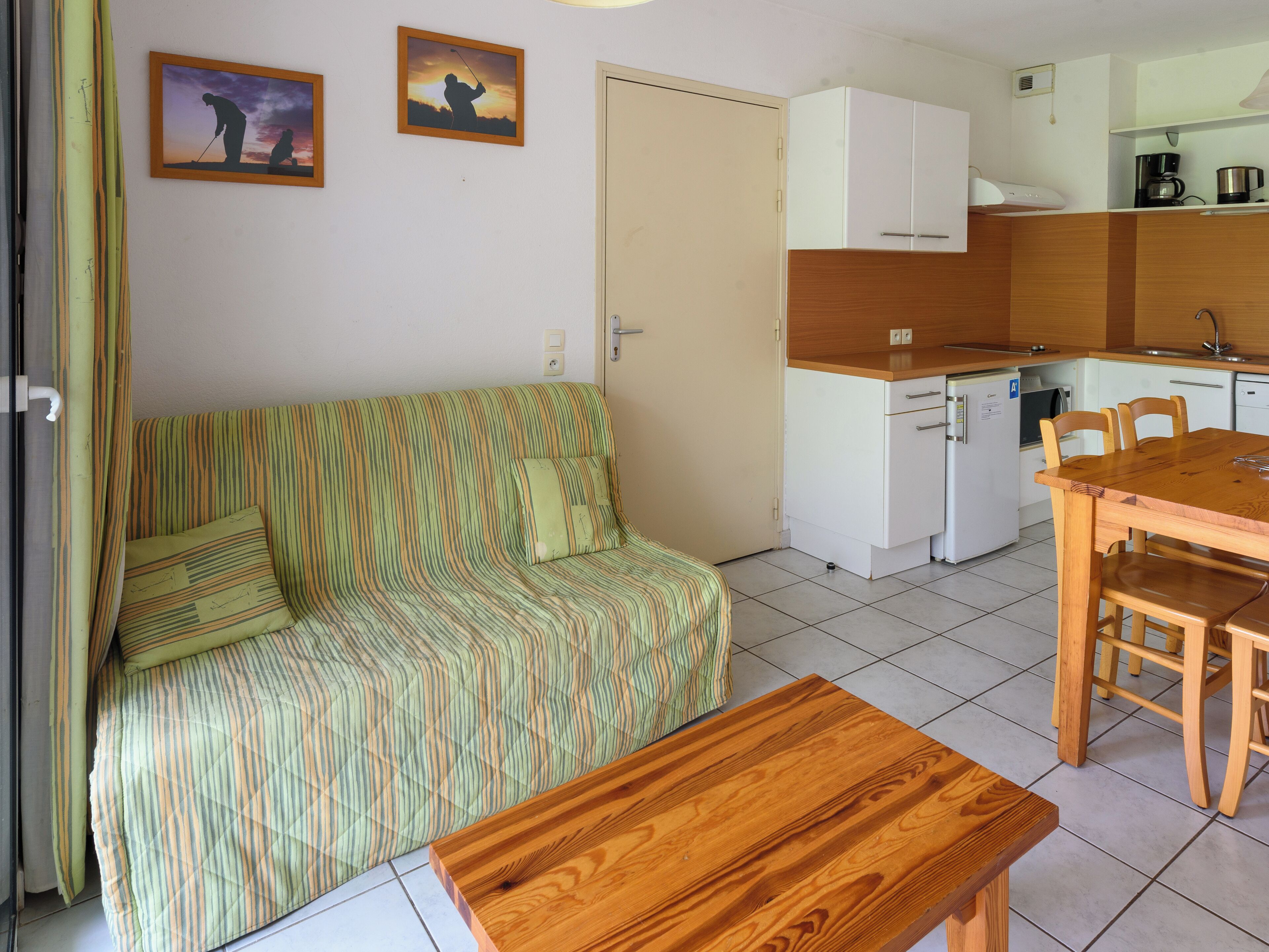 A compact living space with a striped sofa, a wooden dining table, a kitchenette, and two framed pictures on the wall.