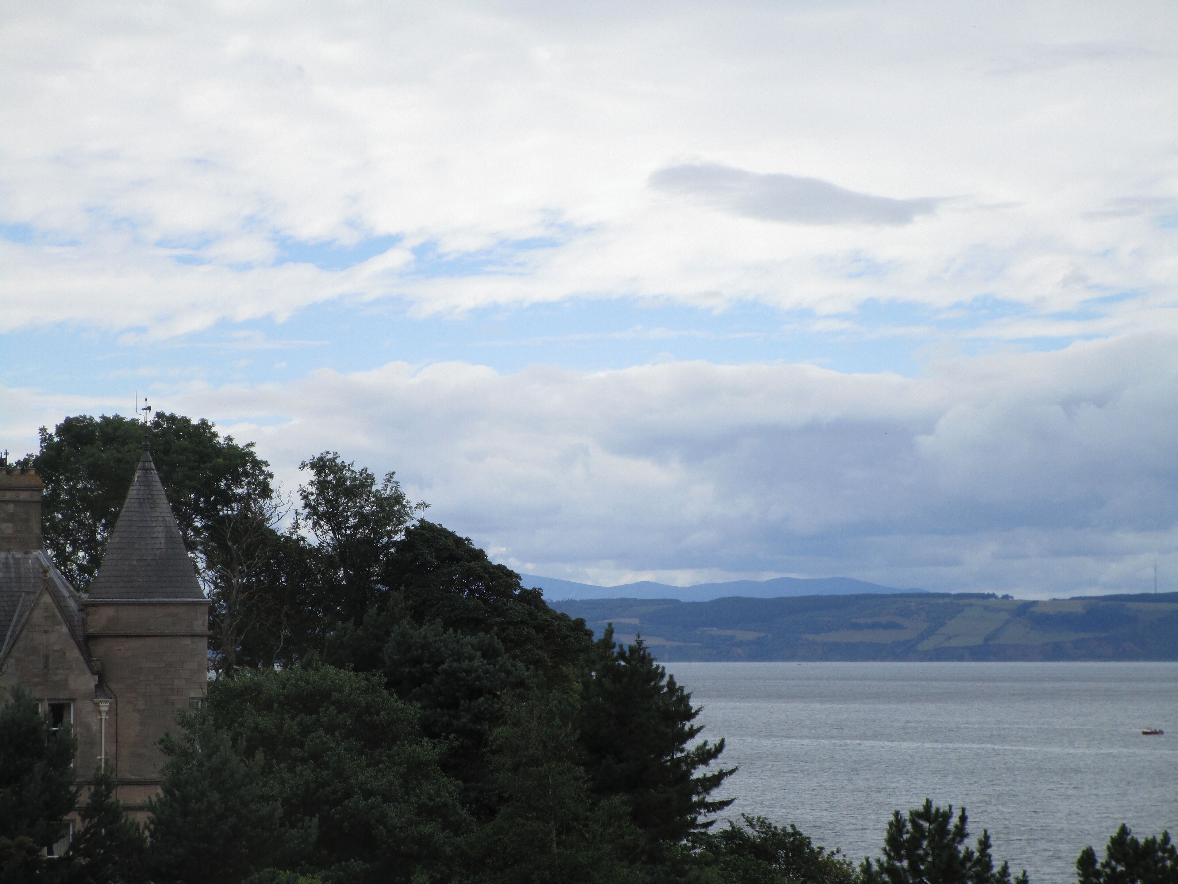 A view of a body of water with a distant shoreline, a stone building with a pointed roof, and a partly cloudy sky.