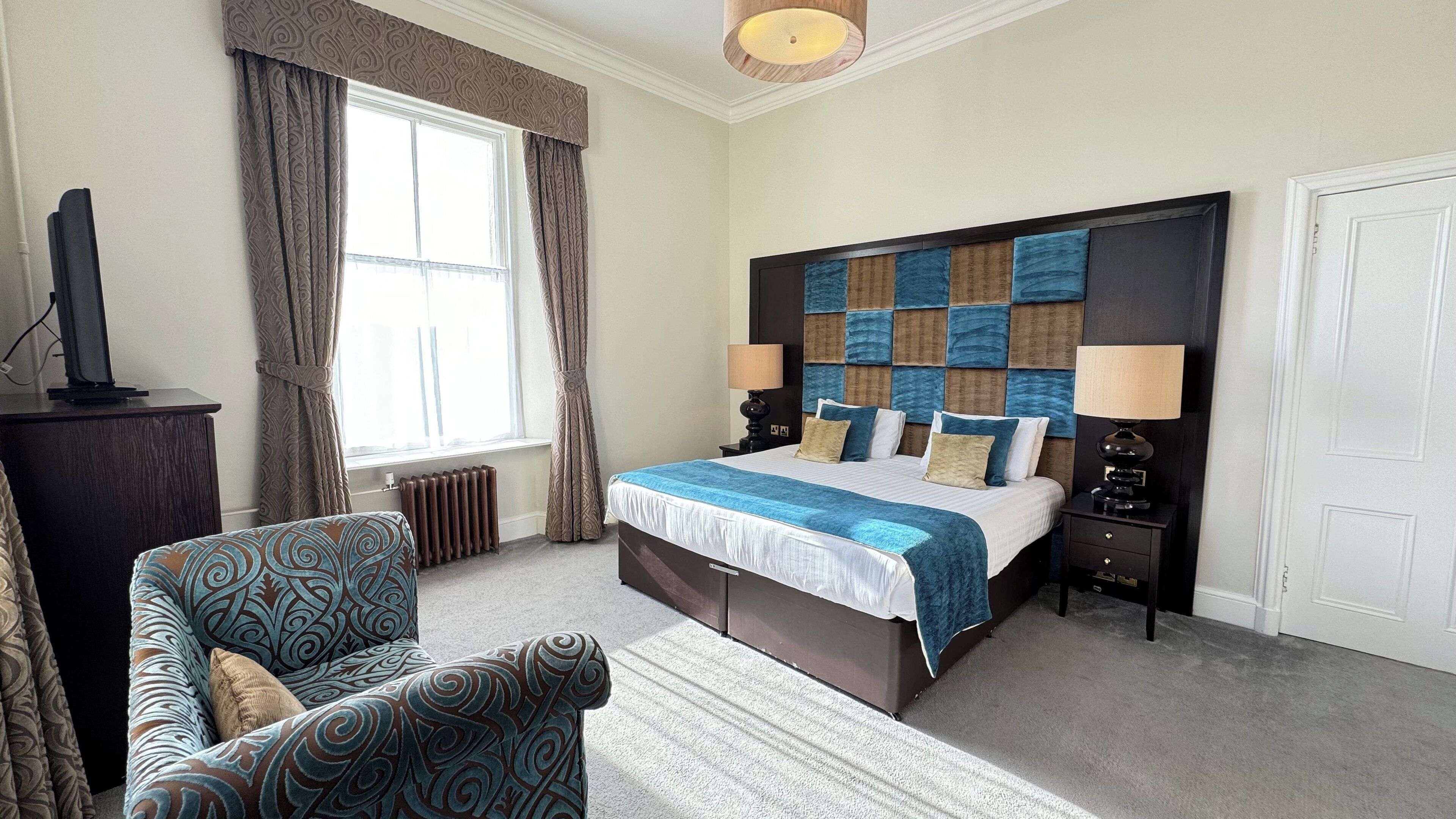 A hotel room with a large bed, a chair, a desk with a computer, and a window with curtains.