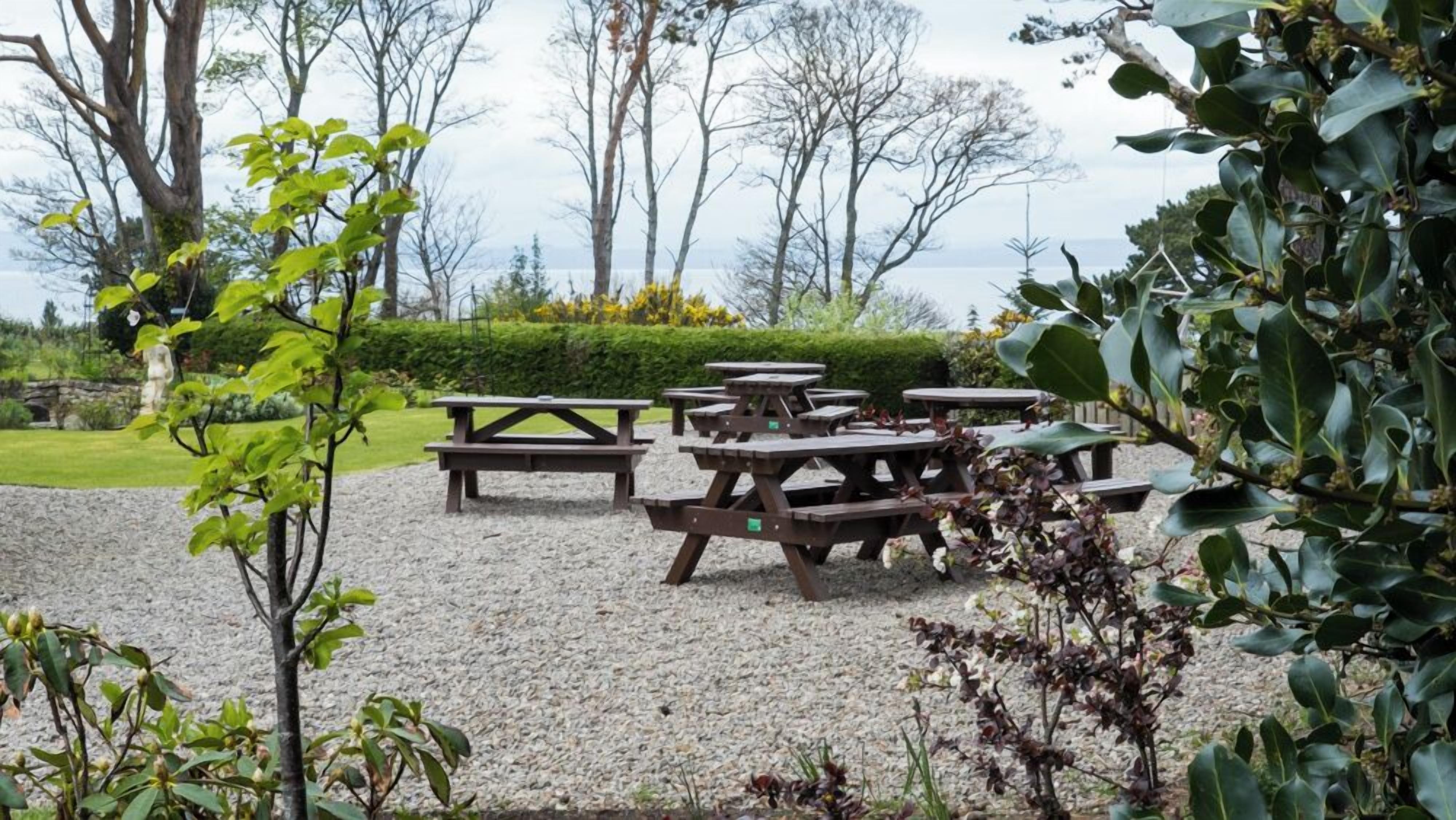 Un área de estar al aire libre con varias mesas de picnic dispuestas sobre grava, rodeada de árboles y un seto.