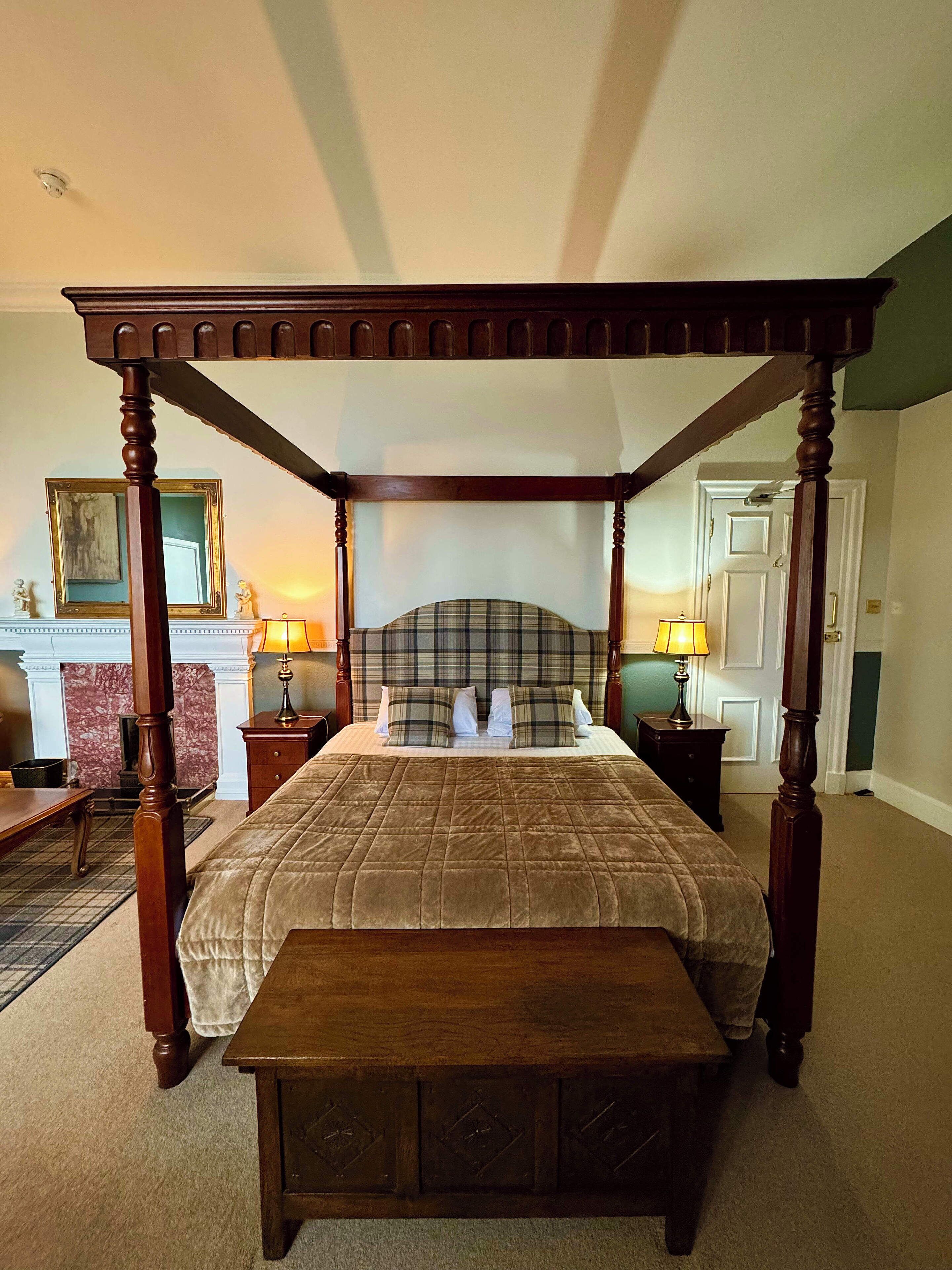 A four-poster bed with a brown quilt, a wooden bench at the foot of the bed, and a fireplace with a marble surround in the background.