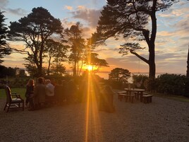 Un grupo de personas reunidas al aire libre al atardecer, con vista al sol poniéndose tras los árboles.
