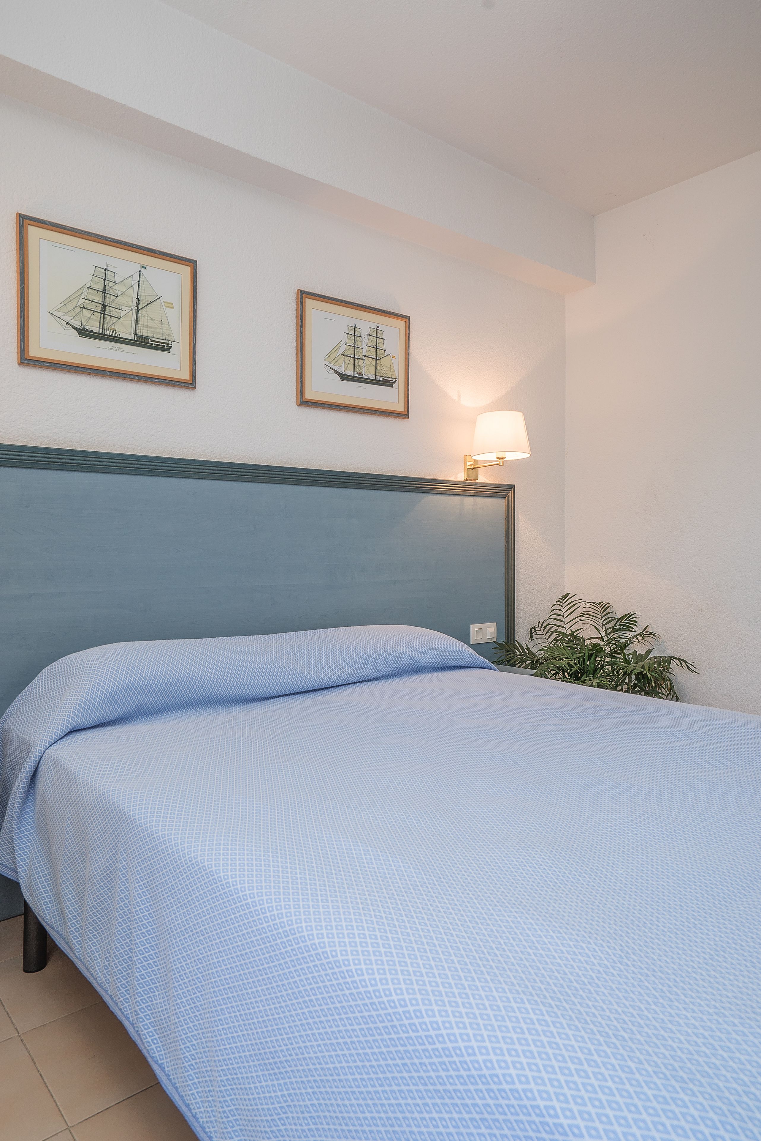 A neatly made bed with a blue bedspread, a headboard, and two framed nautical paintings on the wall.
