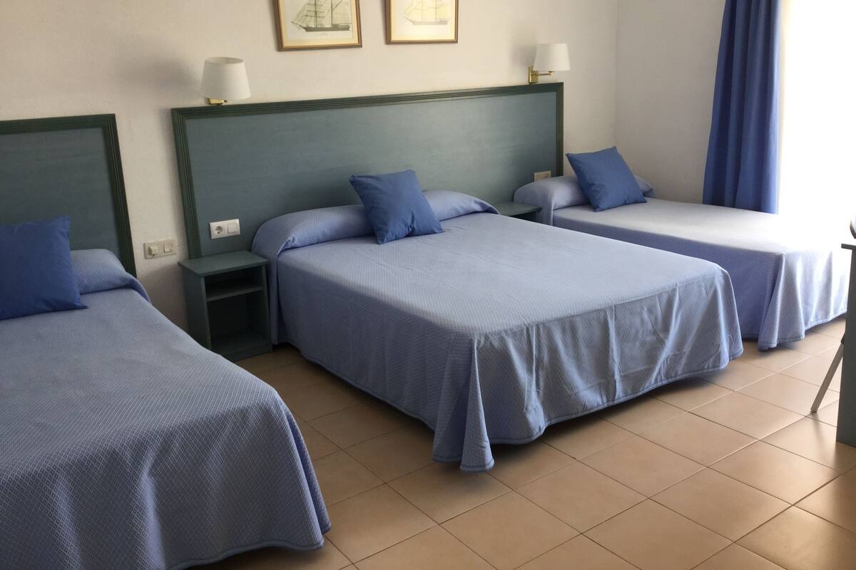 A hotel room with three single beds, each with a blue bedspread and matching pillows. There are two framed pictures on the wall above the beds, a nightstand with a lamp, and a window with blue curtains.