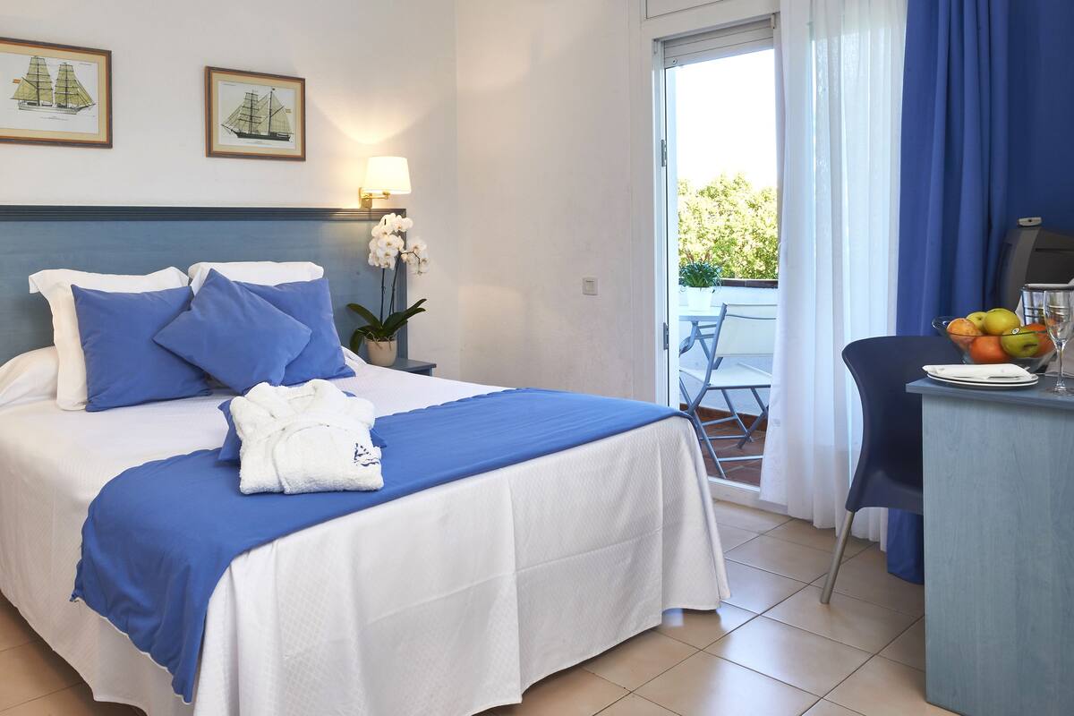 A hotel room with a bed, bedside lamps, a desk with a fruit bowl, and a view of the outdoors.