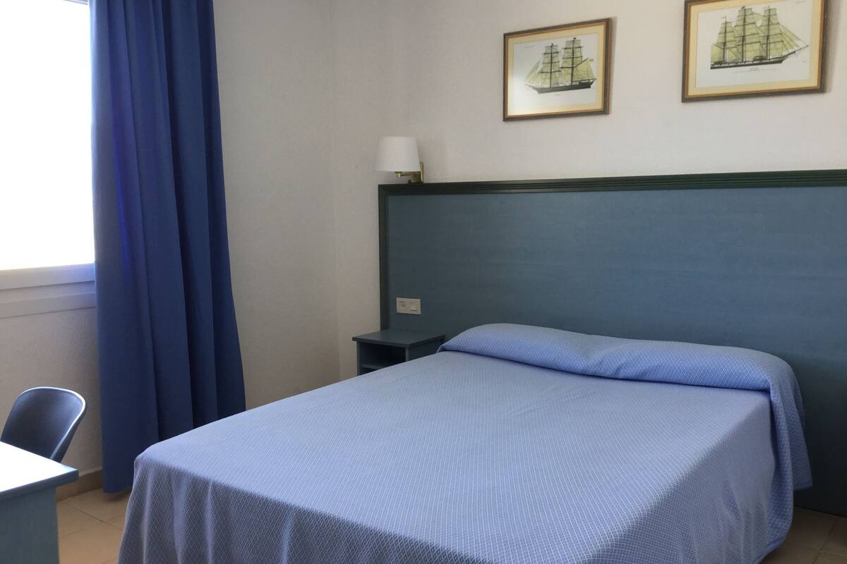 A hotel room with a bed, a desk, a chair, a nightstand, a lamp, and two framed pictures on the wall.