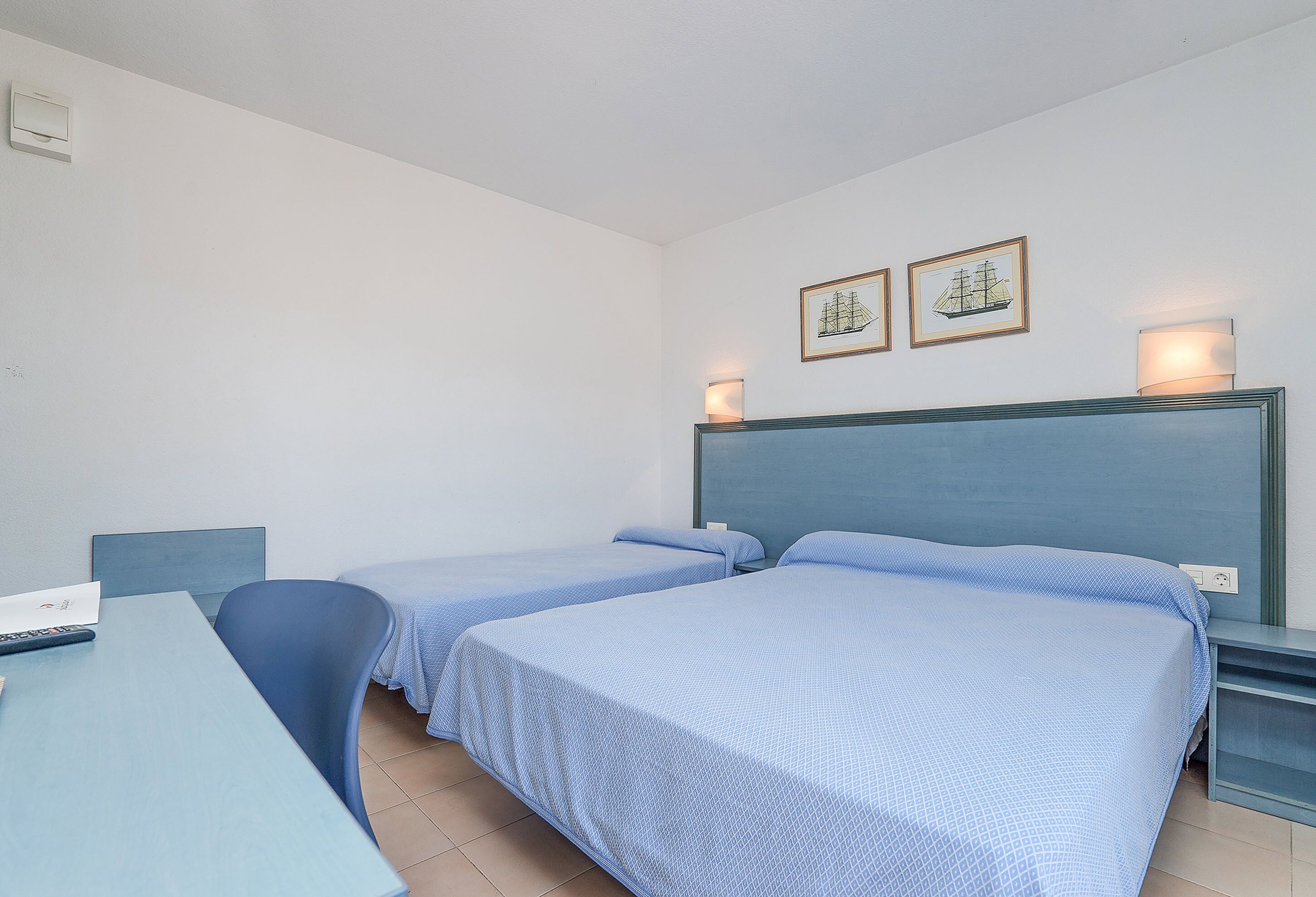Una habitación con dos camas individuales, un escritorio y una silla. Hay dos lámparas de pared y un cuadro enmarcado sobre la cama.