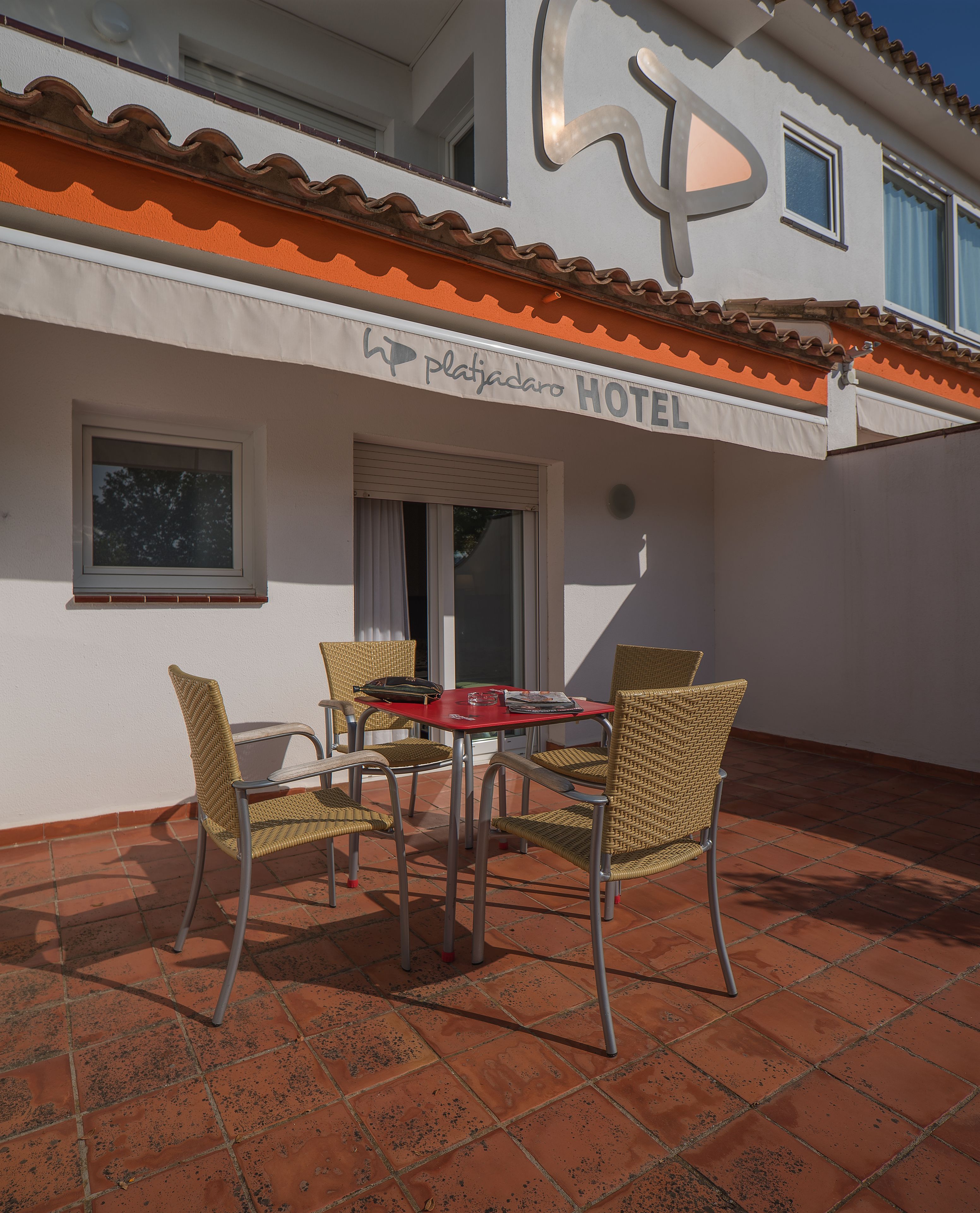 Um pátio de hotel com piso de azulejos, uma mesa com quatro cadeiras de vime e uma placa com a inscrição "Platiacaro Hotel".