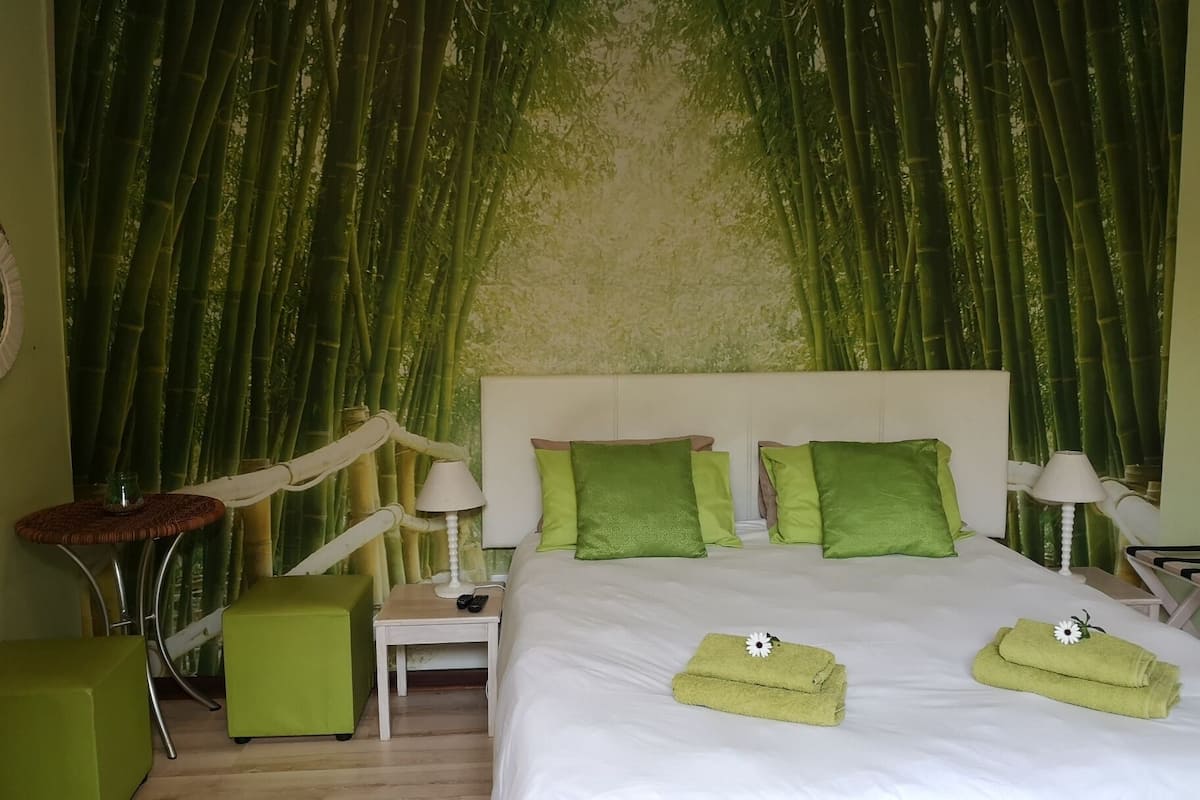 A hotel room with a bed, green pillows, a nightstand with a lamp, a green chair, and a wall with bamboo curtains.