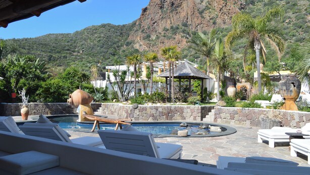A pool area with lounge chairs, a hot tub, and a stone wall. - Hotel Oasi (Lipari)