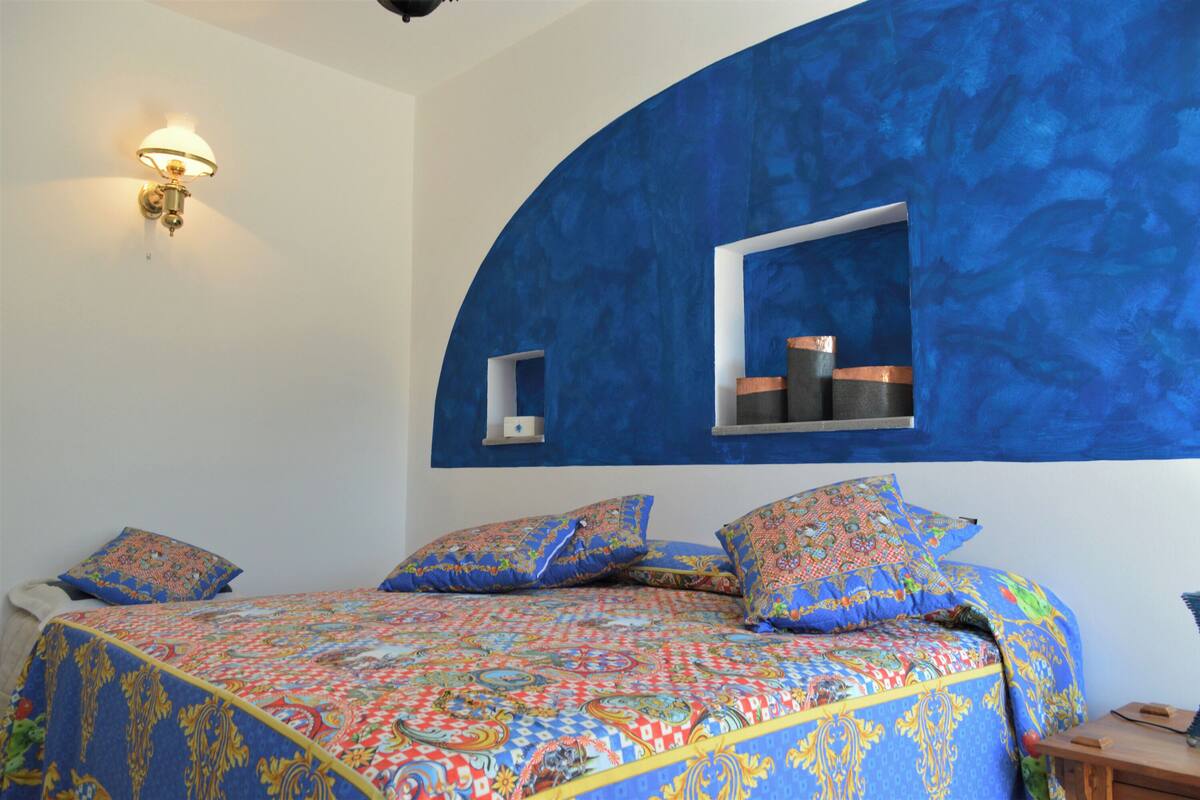 A bedroom with a large bed, blue walls, a window, and a wall-mounted lamp.